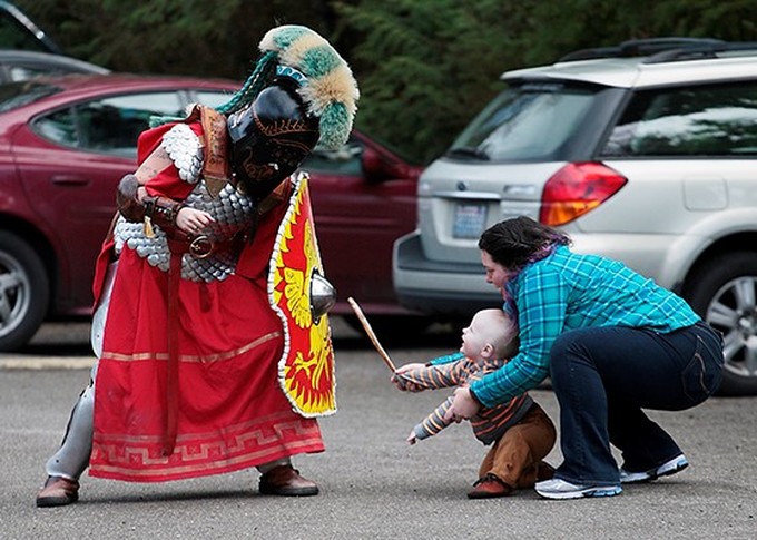 A member of the Myrmidons, dressed in full armor, defends against an "attack" from a toddler (with parent).